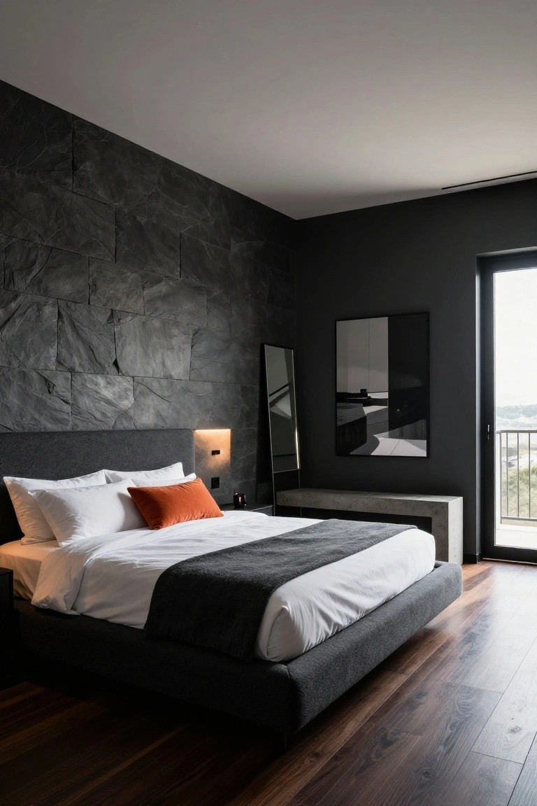Modern bedroom featuring a dark textured stone accent wall behind a gray upholstered platform bed with white sheets, orange pillow, gray throw, full-length mirror, and glass balcony door overlooking landscape.