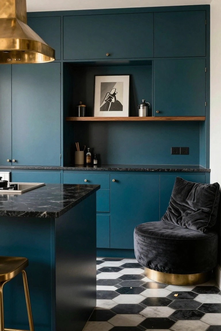 Compact modern kitchen with teal cabinets, black marble island countertop, gold range hood, black velvet pouf chair beside the island, wood shelves, and black-and-white hexagonal tile floor.