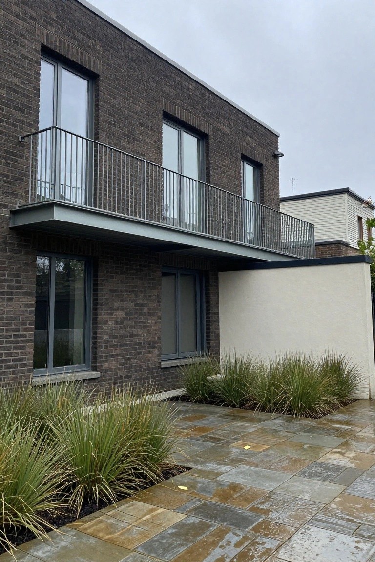 Modern two-story house exterior with dark brick walls, a protruding gray metal balcony with slim railings, large gray-framed windows, a white rendered wall panel, ornamental grasses in beds, and wet stone paving under cloudy skies.