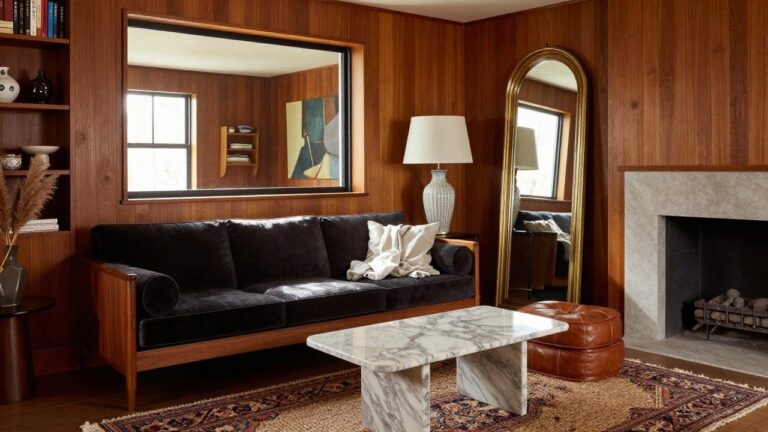 Tall gold-framed arched floor mirror leaning against wood-paneled wall in a living room with gray sofa, leather chairs, patterned rug, marble coffee table, pampas grass arrangement by stone fireplace, and abstract wall art.