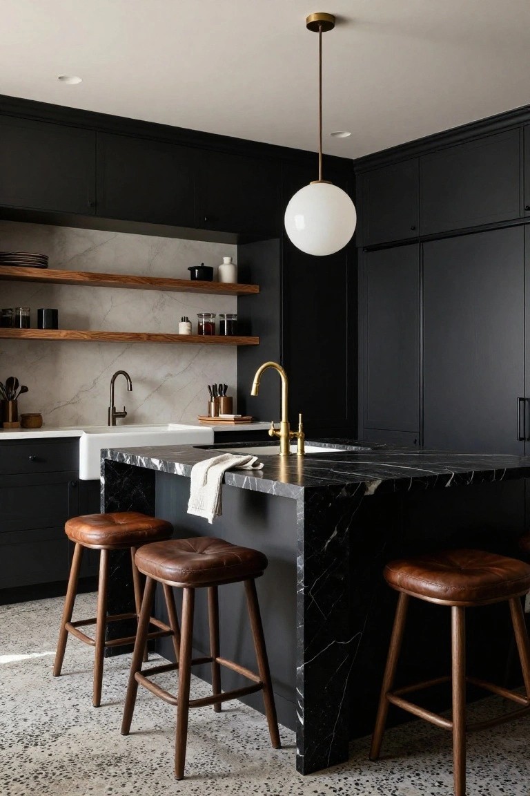 Kitchen interior with black cabinets, white farmhouse sink, black marble-topped island, brass faucet, wood open shelves, and brown leather bar stools on a terrazzo floor.