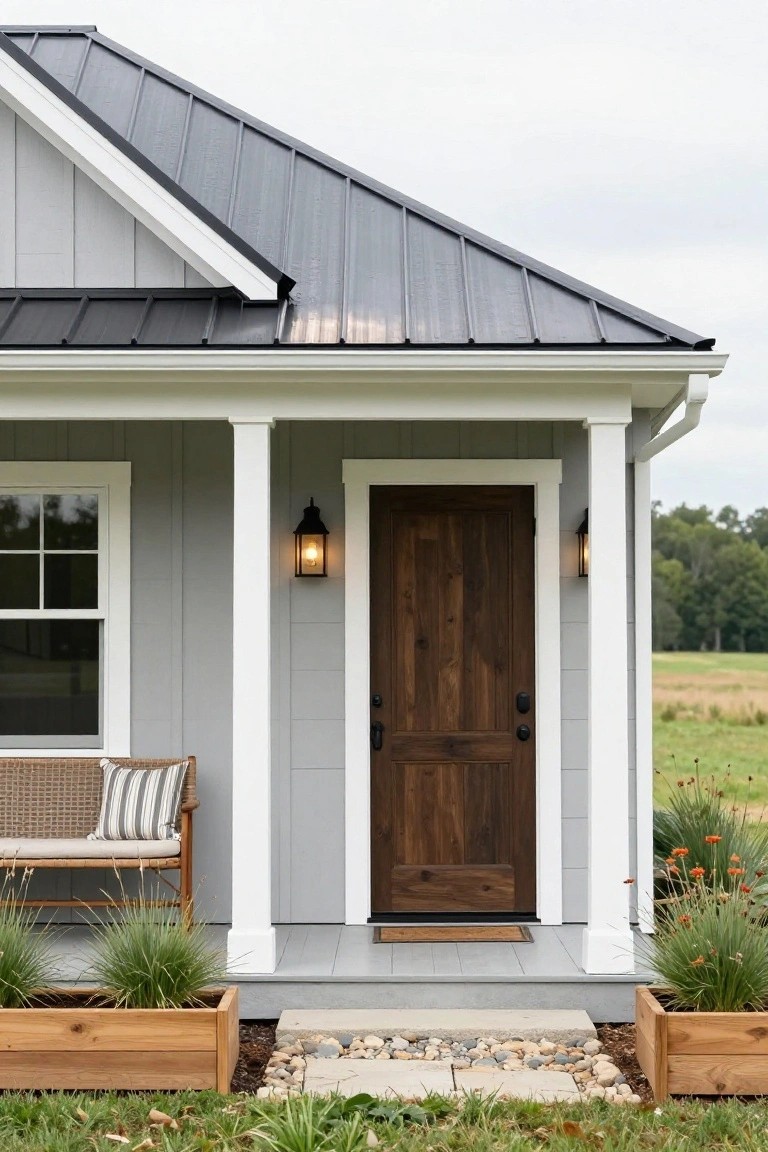 Close view of a light gray clapboard house exterior with black metal roof, white porch columns supporting the overhang, dark wood front door with lanterns, wicker bench, wooden planters, and gravel path on a lawn with trees in background.
