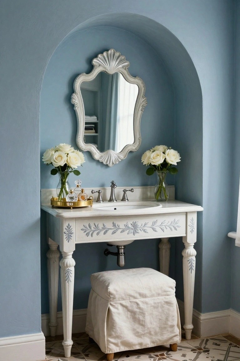 Blue-painted arched wall recess with white scalloped mirror, white vanity table holding sink and gold tray of perfume bottles, flanked by vases of white roses, plus a skirted white stool below.