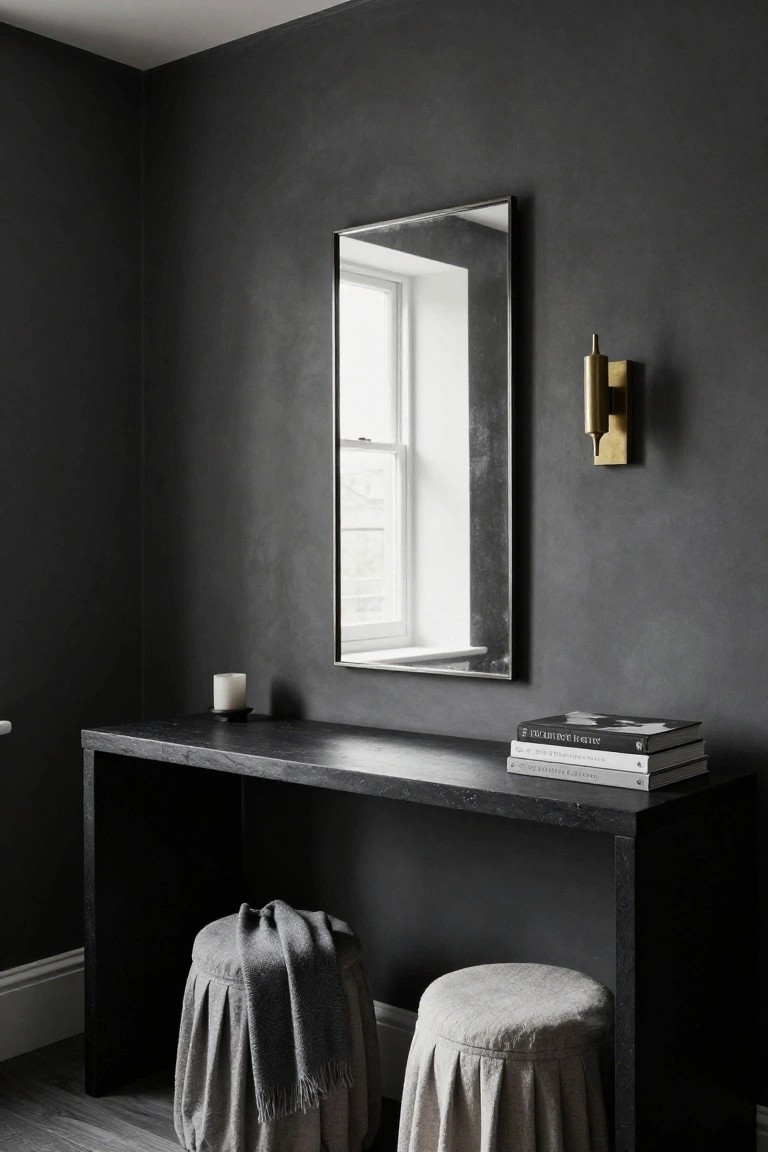 Tall rectangular mirror above a black stone console table with two fabric-covered stools nearby, gold wall sconce, candle, and books on the table in a dark gray room with a window.