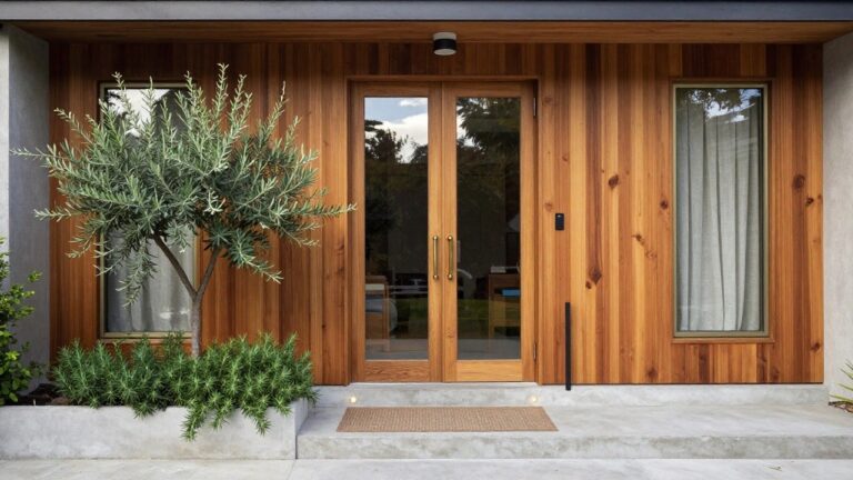 Front entrance of a house with vertical cedar wood siding, a tall clear glass door with brass handle and lock, an olive tree and potted rosemary plant to the left, beige doormat, and concrete steps.