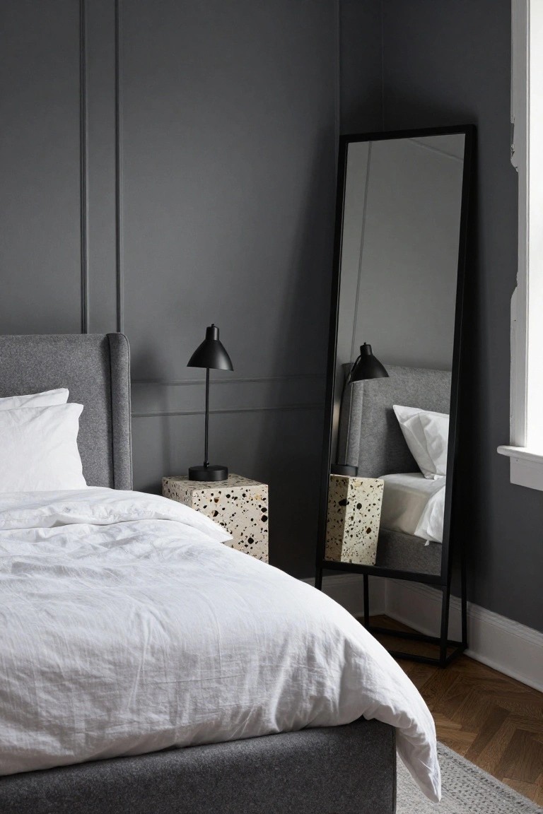 Corner of a bedroom with dark gray paneled walls, gray upholstered bed with white linens and pillows, terrazzo cube side table holding a black lamp, tall black-framed full-length mirror, and herringbone wood floors.