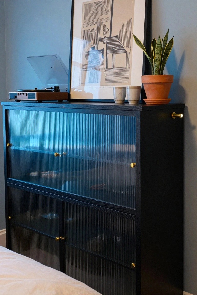 Tall black cabinet with blue-tinted fluted glass doors and gold knobs against a blue wall, topped with a wood record player, two ceramic mugs, a snake plant in terracotta pot, and framed abstract art nearby, with a bed edge visible at bottom.