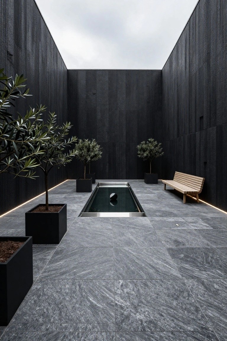 Tall black wooden walls enclose a narrow rectangular reflecting pool in a courtyard, flanked by potted olive trees and a wooden bench on gray slate flooring with LED edge lighting.