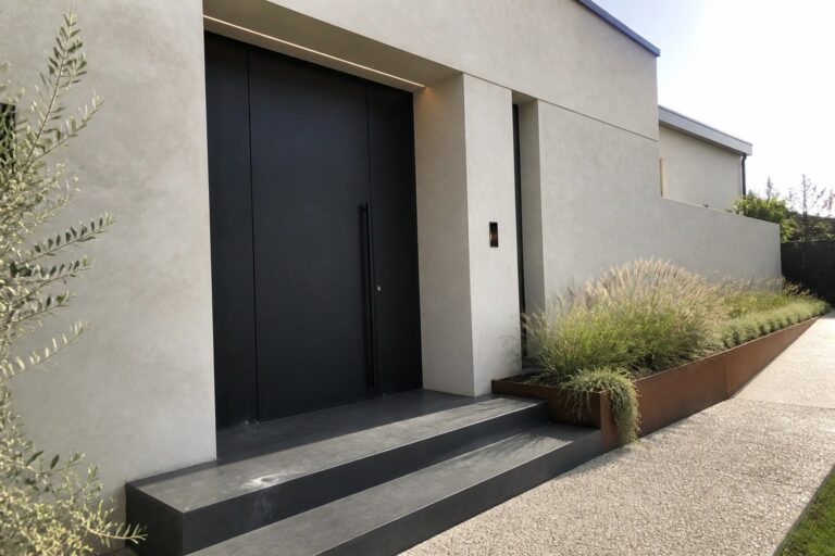 A dark flush-mounted door set into a light concrete exterior wall with dark steps leading up to the entrance.