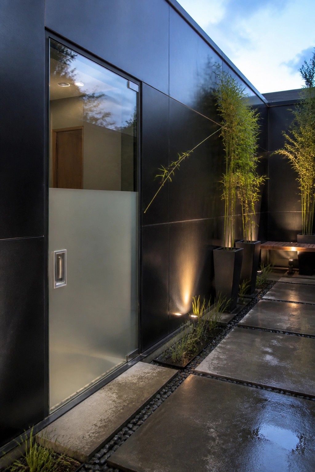 Modern black exterior wall with a large glass entry door featuring a frosted lower panel set flush into the wall, next to tall bamboo plants in black planters and wet stone pavers.
