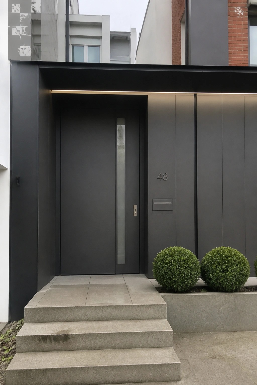 A dark gray modern entry door with a vertical glass panel is set flush into a matching wall, with concrete steps leading up and two round shrubs to the right.