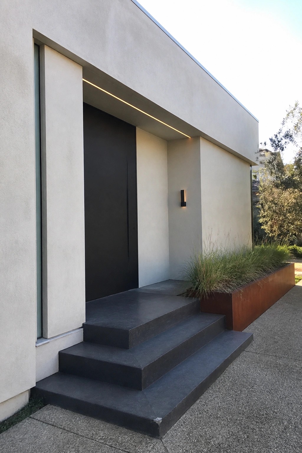 A dark flush-mounted door set into a light concrete exterior wall with dark steps leading up to the entrance.
