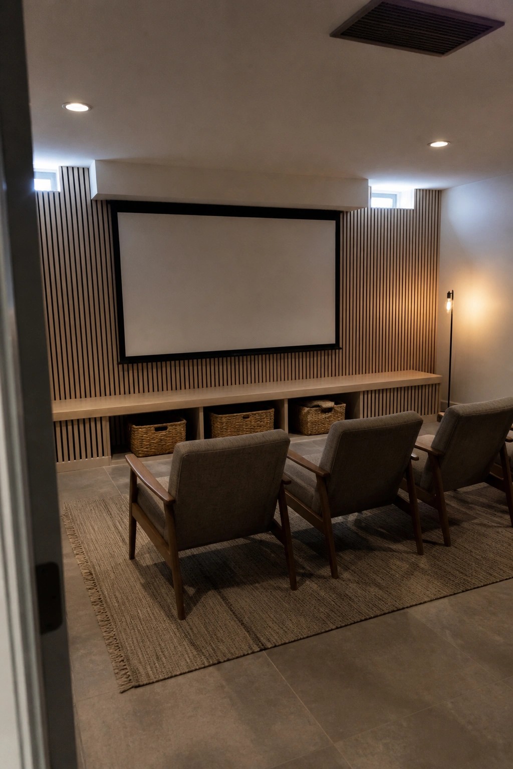 A home theater room showing a large projection screen on a wall covered in vertical wood slats, with three upholstered chairs on a rug and woven baskets stored under a long built-in shelf.