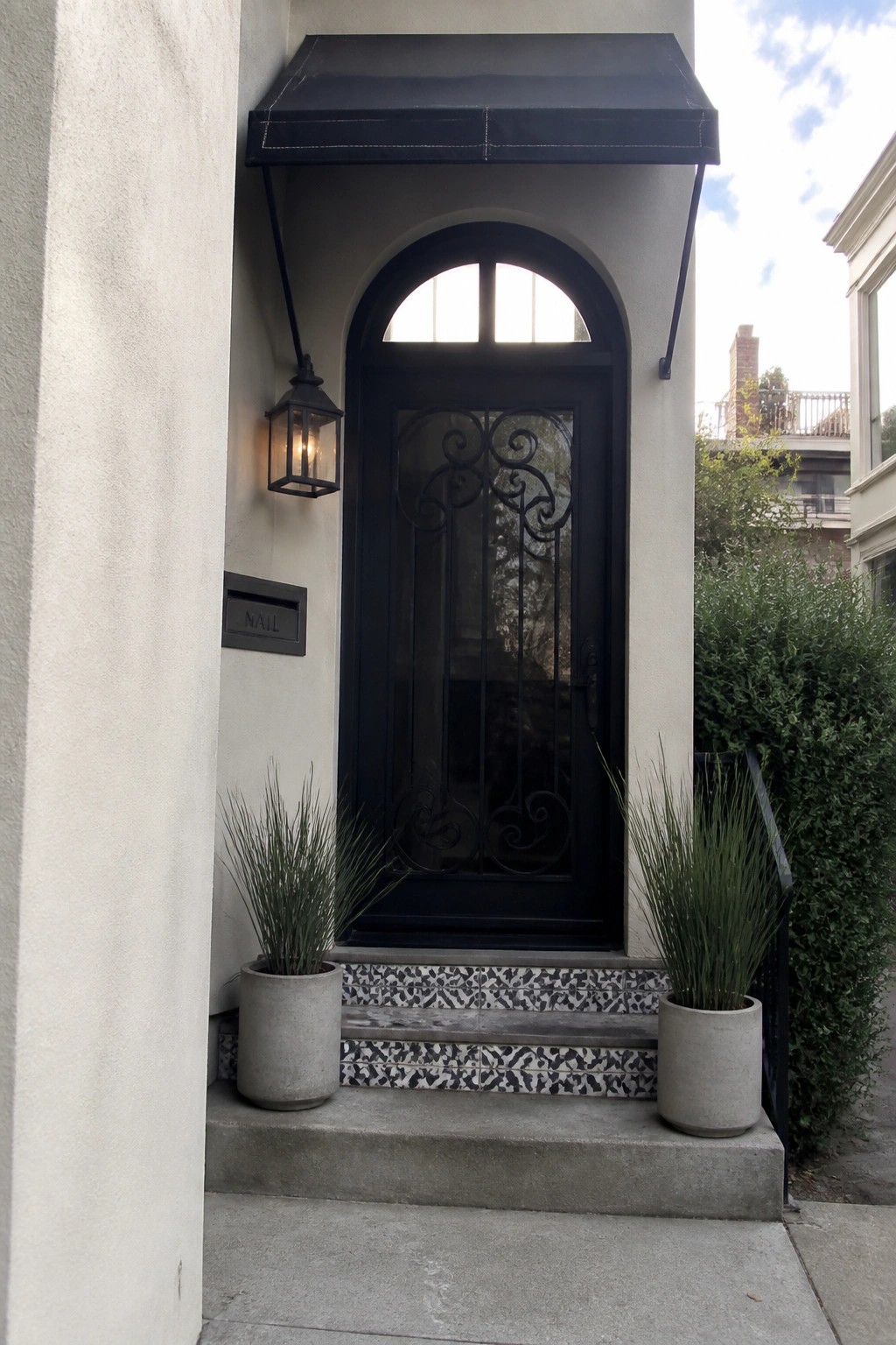 Black arched front door with decorative ironwork, black awning above, wall lantern to the left, and two potted plants on the steps.