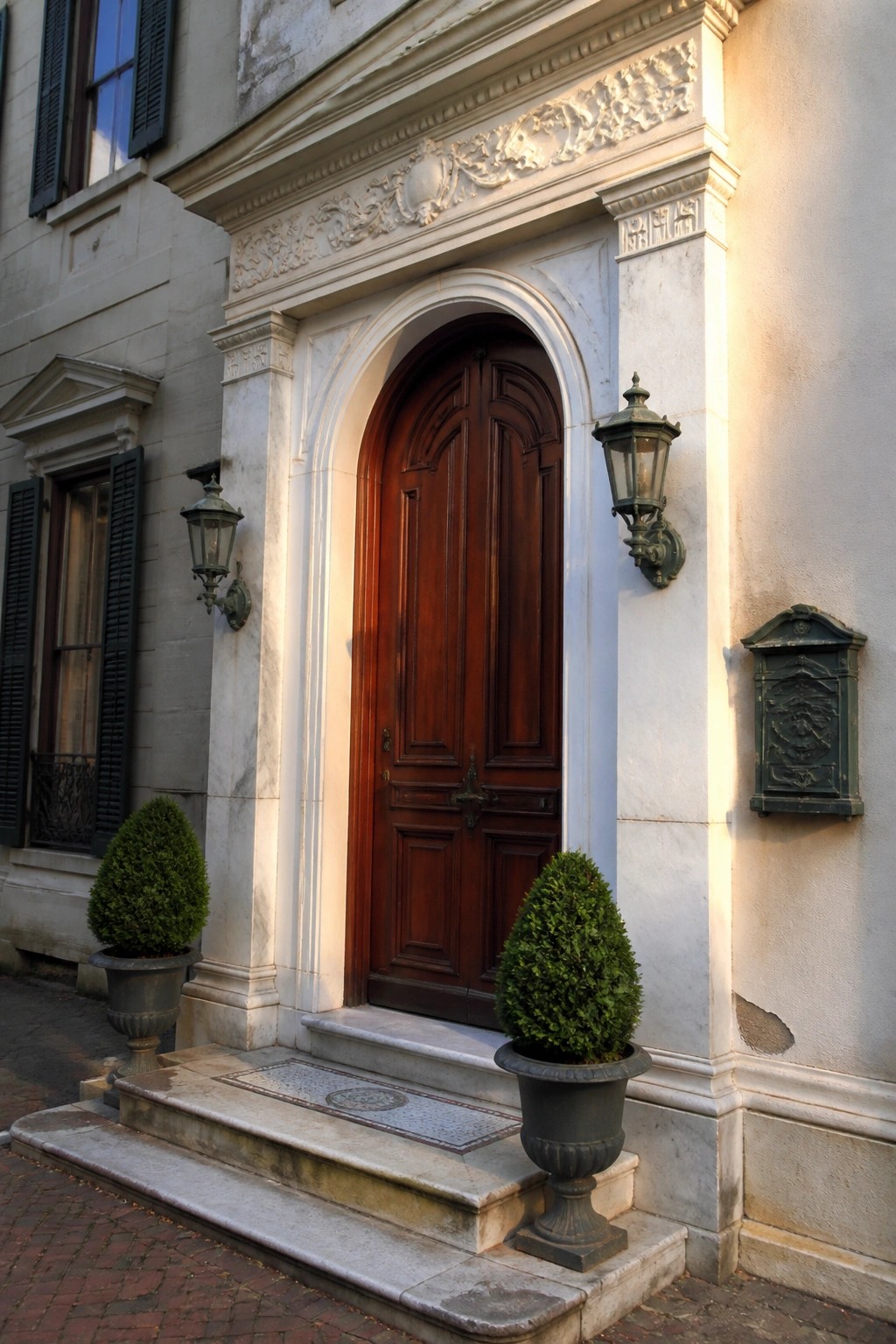 A dark wooden double door centered in an arched marble surround, with wall lanterns on both sides and two potted shrubs on the stone steps.