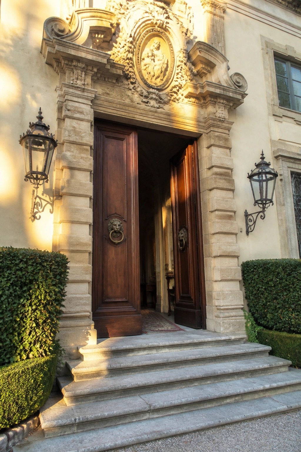 Ornate double wooden doors stand open beneath a carved stone surround with a central medallion, flanked by wall lanterns and leading up from stone steps.