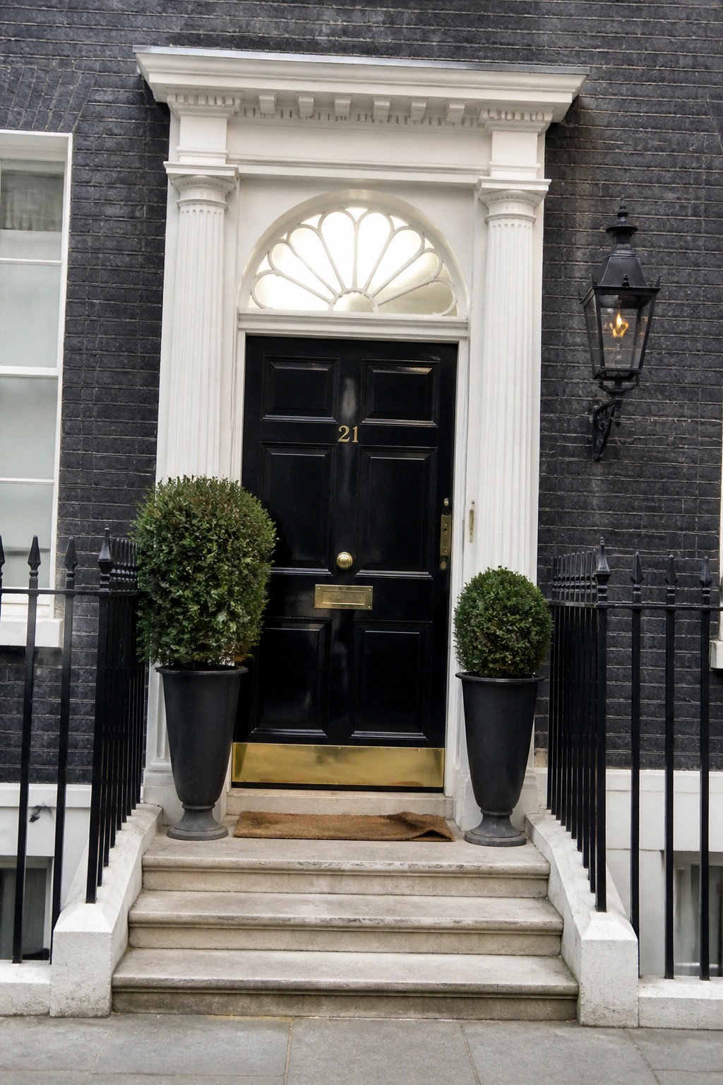 A black six-panel front door with a brass kickplate sits beneath a white arched fanlight, framed by two white fluted columns, with black planters holding shrubs on the stone steps.