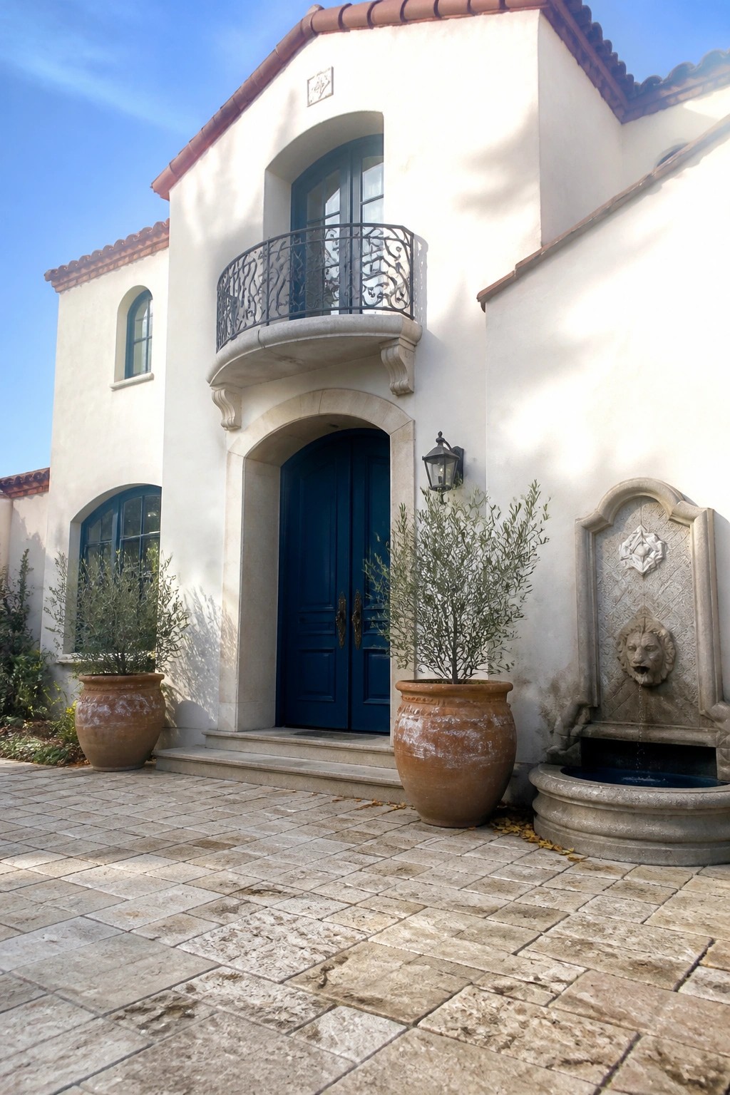 White stucco villa entrance with blue doors, balcony, potted trees, and stone lion fountain