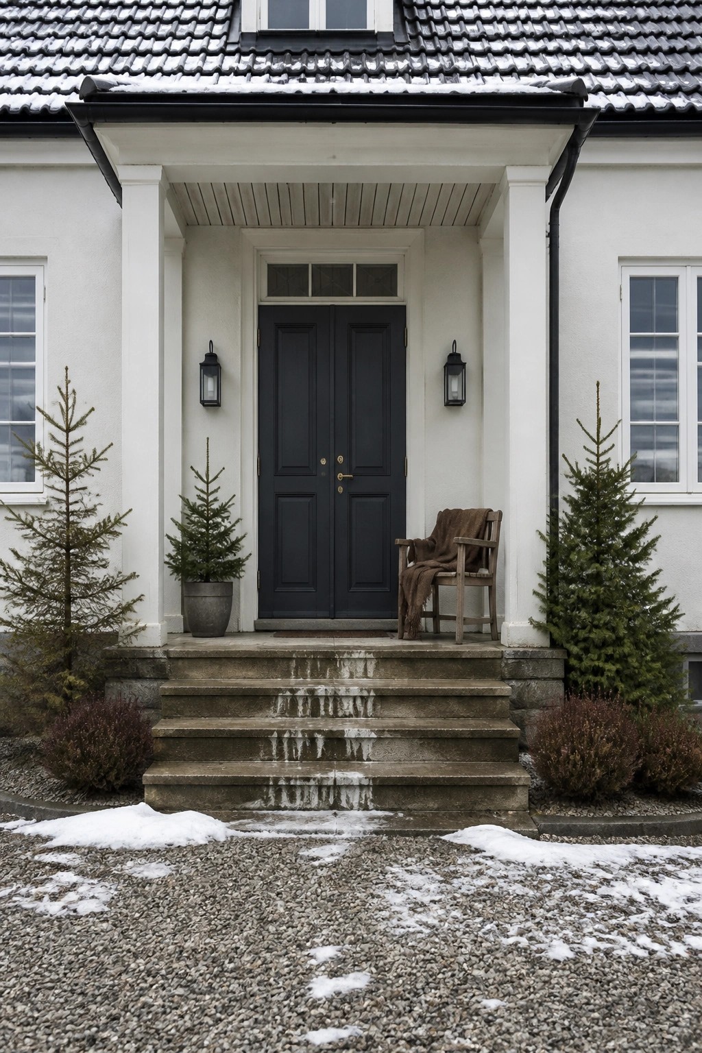 A white house exterior with a dark double door centered under a small portico, flanked by wall lanterns and potted evergreens, with stone steps leading to a gravel driveway.