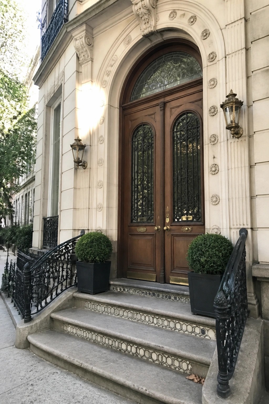 Ornate wooden double doors with arched tops, black iron grilles, and a stained glass transom above, set in a light stone facade with steps and potted shrubs.