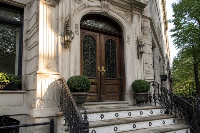 19 Ornate Luxury Doors Inspired by European Architecture Ornate wooden double doors with arched tops, black iron grilles, and a stained glass transom above, set in a light stone facade with steps and potted shrubs.