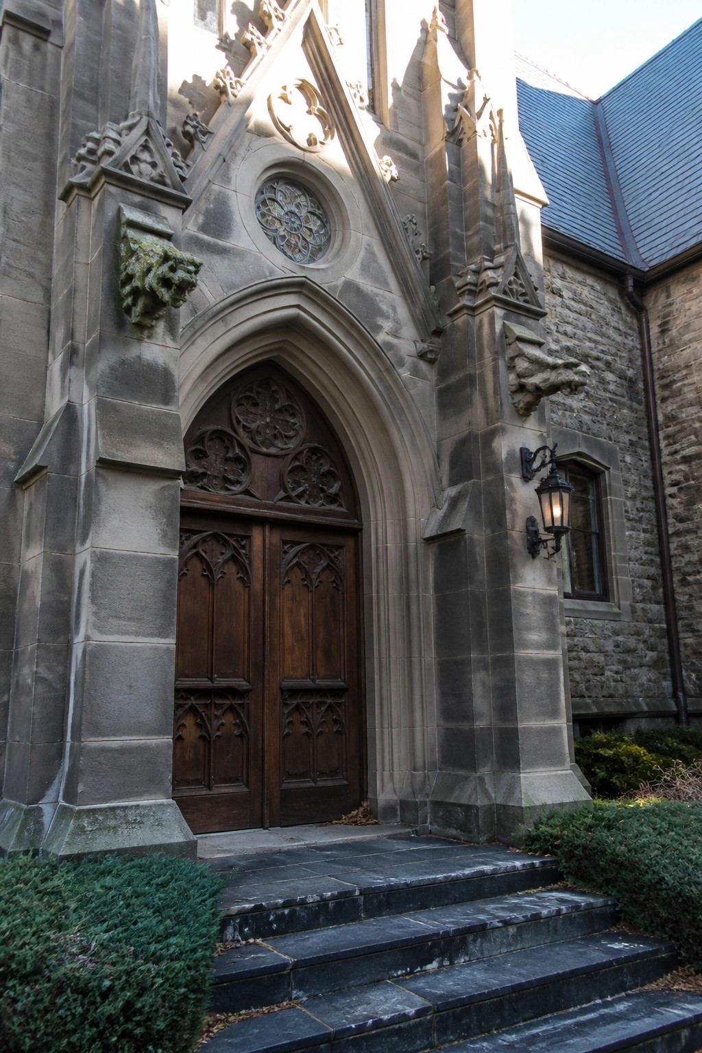 A stone facade with double dark wood doors set under a tall pointed arch, a circular stained glass window above, and stone steps leading to the entrance.