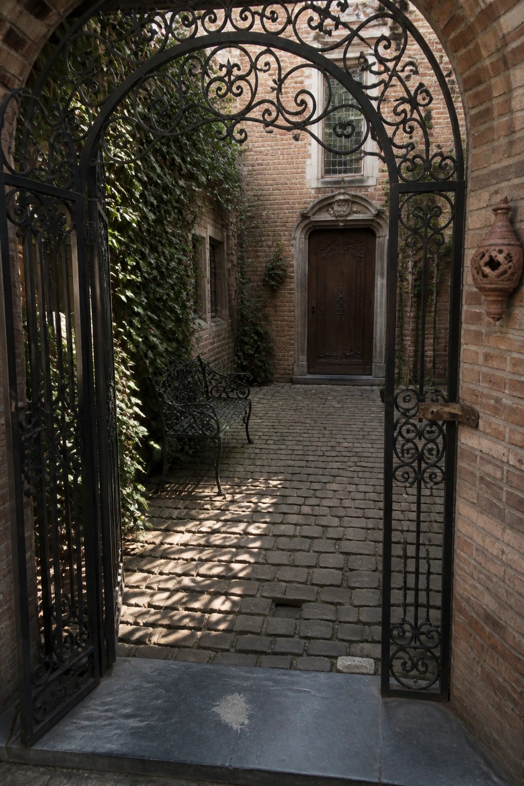 An open ornate black wrought iron gate frames a view into a brick courtyard with a cobblestone path leading to a wooden door.