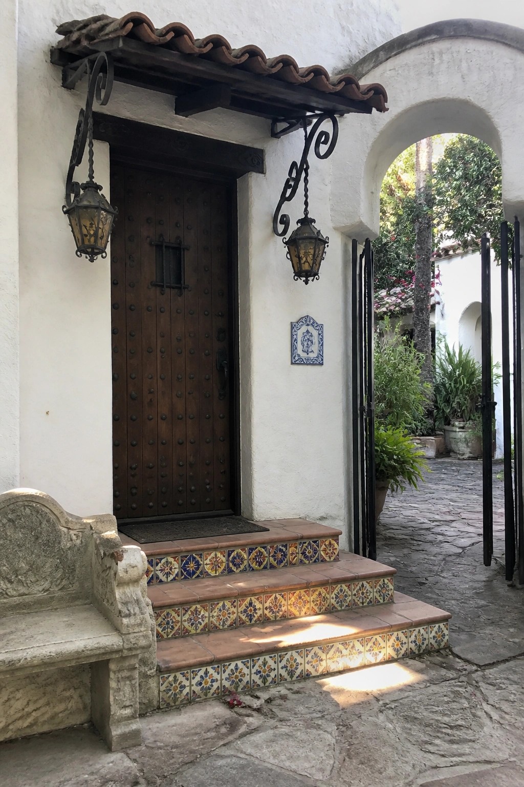 A dark wooden door with metal studs flanked by two hanging lanterns, set in a white stucco wall above colorful tiled steps.