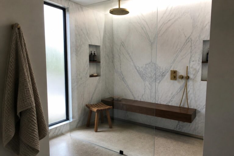 19 Resort-Style Luxury Shower Ideas Featuring Hotel-Inspired Rainfall Systems A modern walk-in shower with white marble walls in a chevron pattern, two recessed niches holding toiletries, a rainfall showerhead, a wooden bench, and a glass door.