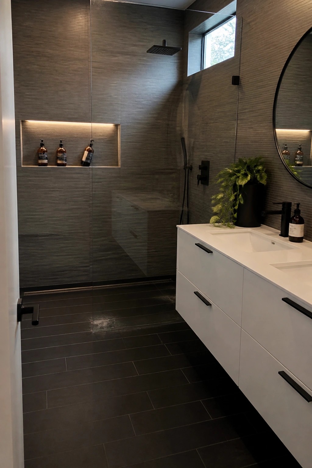 A modern bathroom interior showing a glass-enclosed shower with dark gray textured tiles, a linear floor drain, a recessed lit niche with three bottles, and a white double vanity with black fixtures.