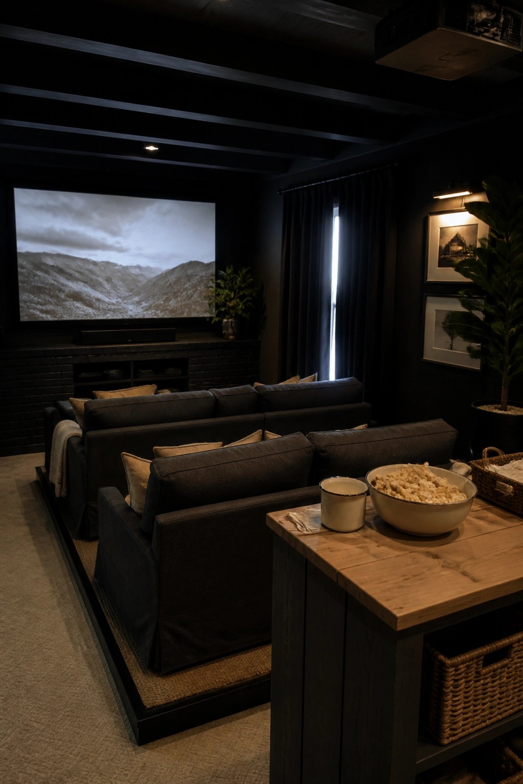 A home theater room with black walls and ceiling, a large screen showing a landscape, dark sectional sofas, and a wooden table with popcorn in the foreground.