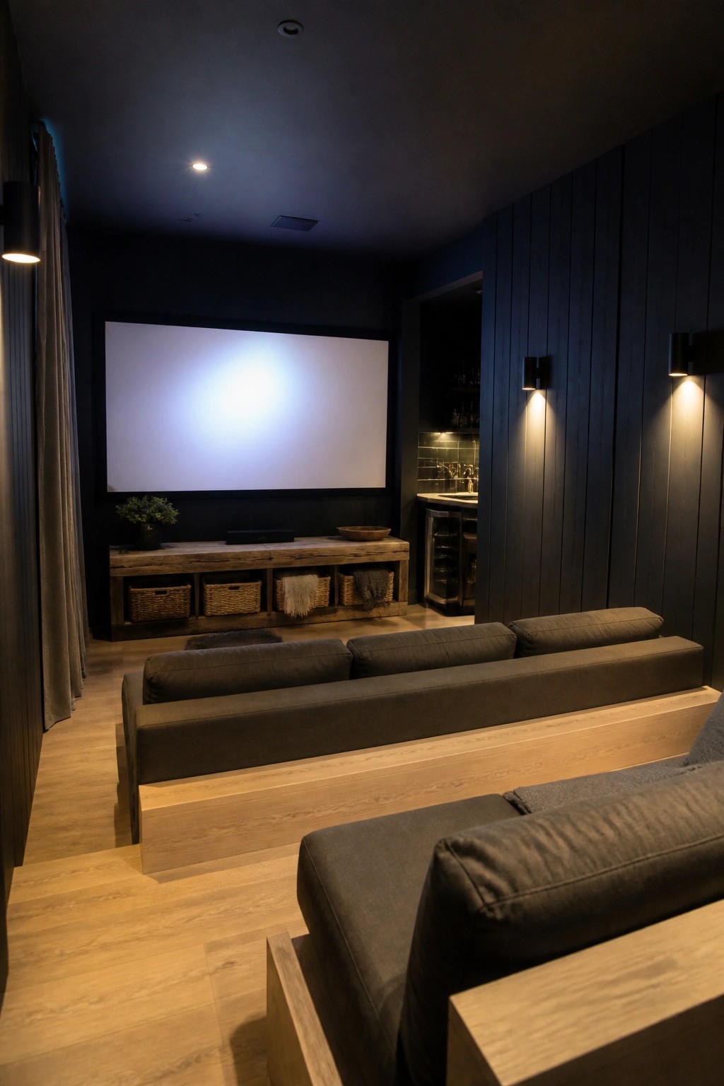 A dimly lit home theater with black walls, a large blank screen on the back wall, and three rows of dark gray sofas arranged on light wood tiered platforms.