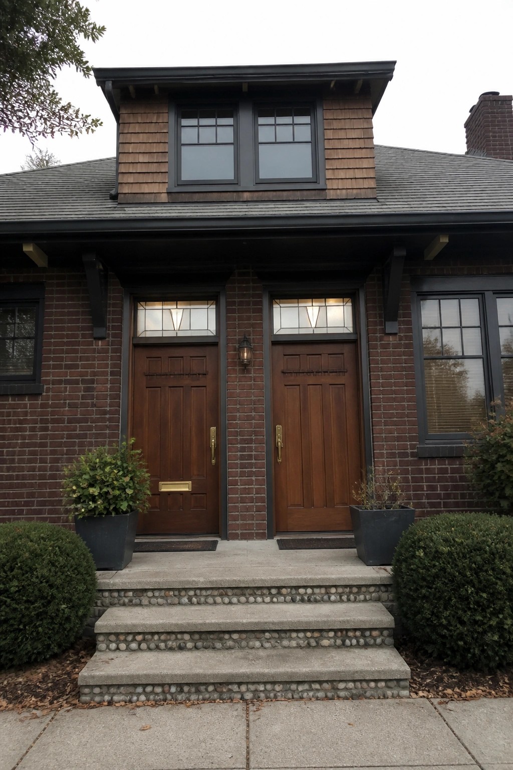 Front view of a brick house with two matching brown wooden doors side by side, each topped by a divided glass transom, concrete steps with pebble edging, and potted plants at the base.