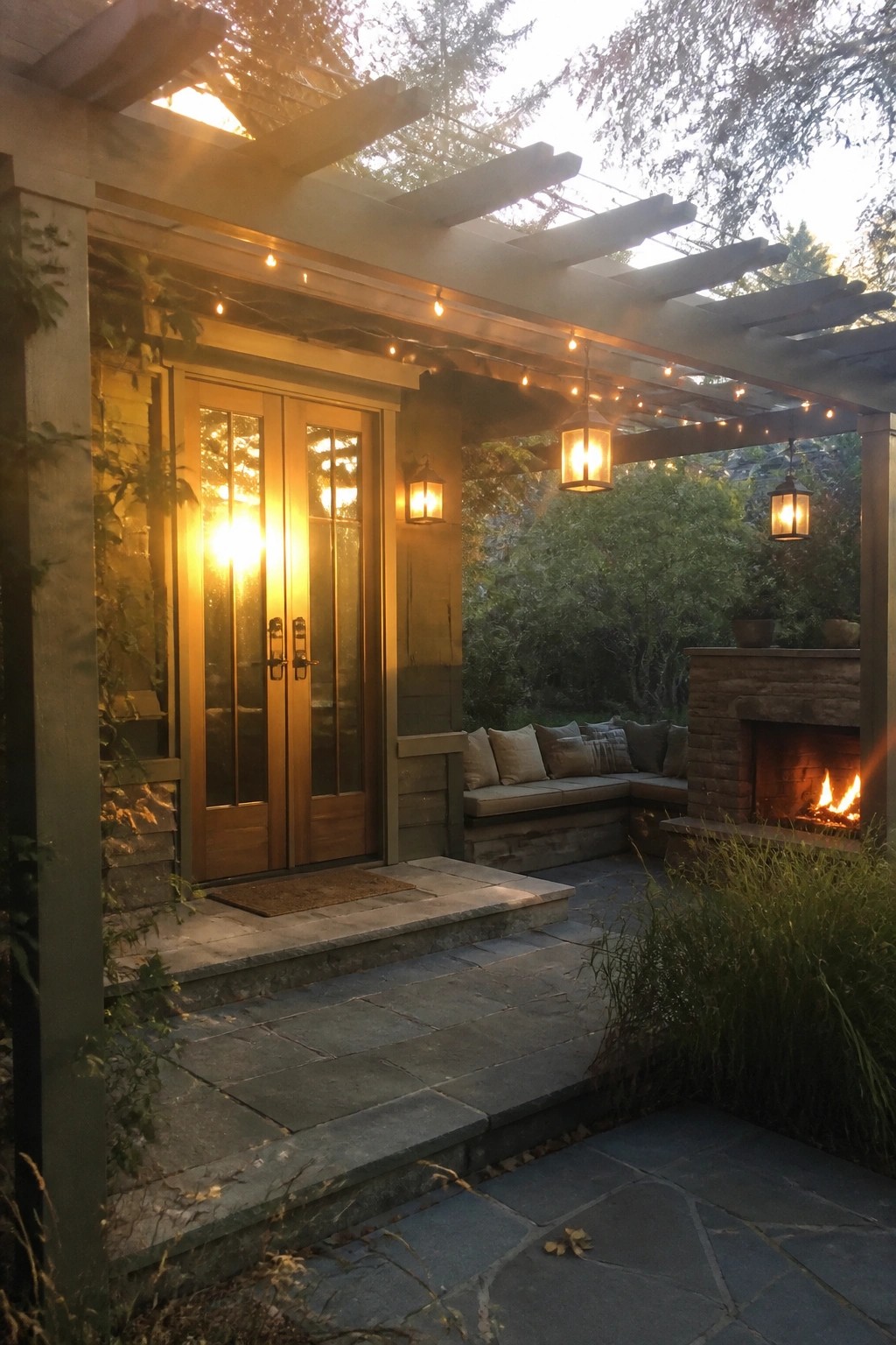 Wooden double doors with glass panels stand under a pergola strung with lights beside an outdoor seating area and stone fireplace.