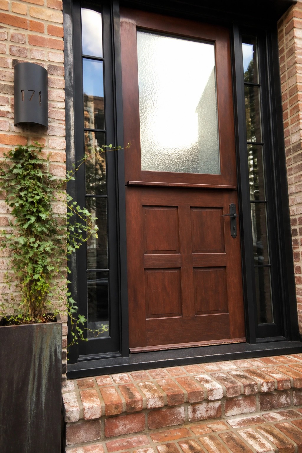 A dark wood front door with four lower panels and a textured glass upper panel, framed in black with matching glass sidelights on a red brick exterior.