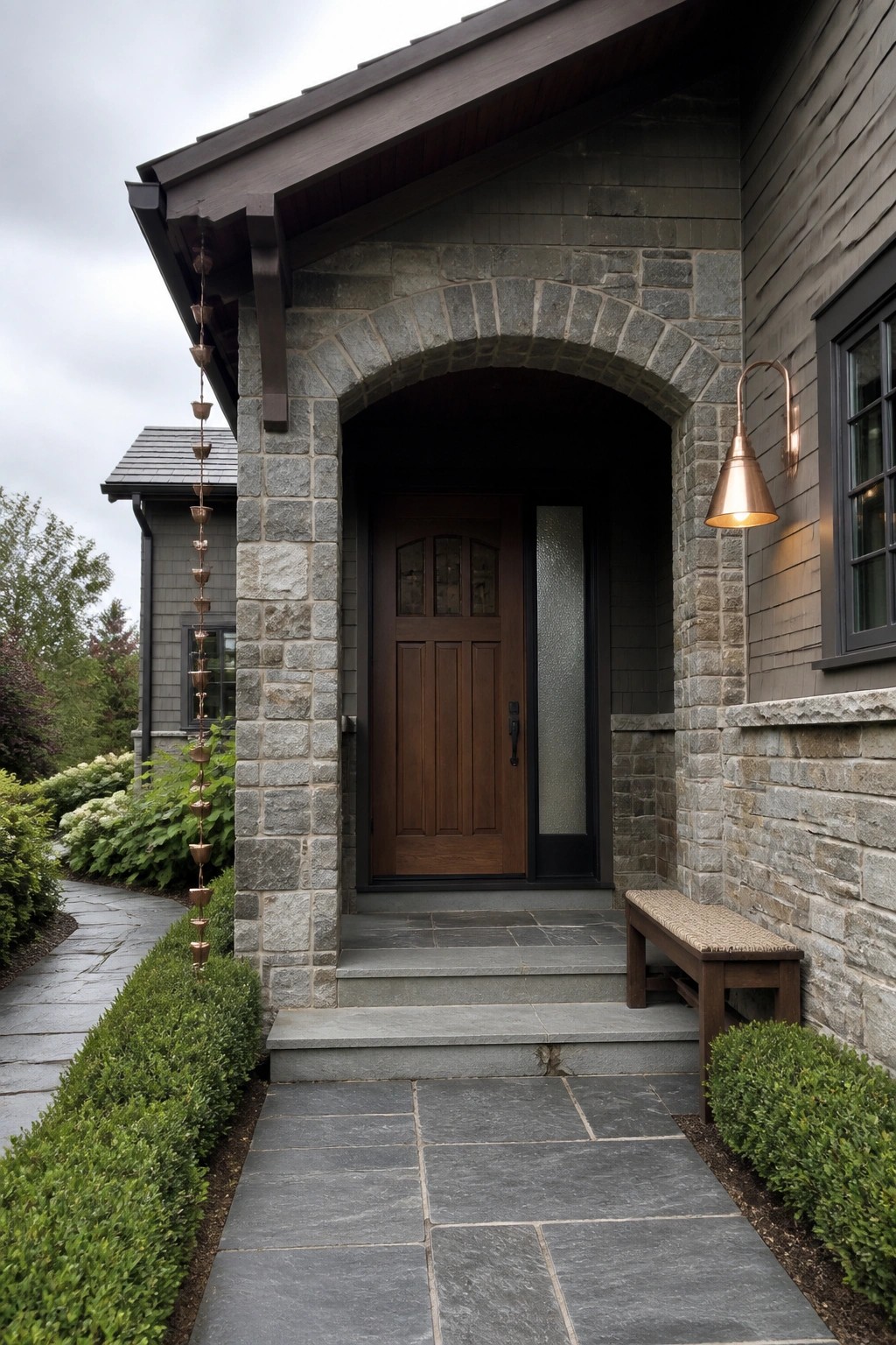 A Craftsman-style house entrance featuring a dark wood door beneath a gray stone arch, with stone pillars, a copper wall sconce, and a stone pathway leading to the steps.
