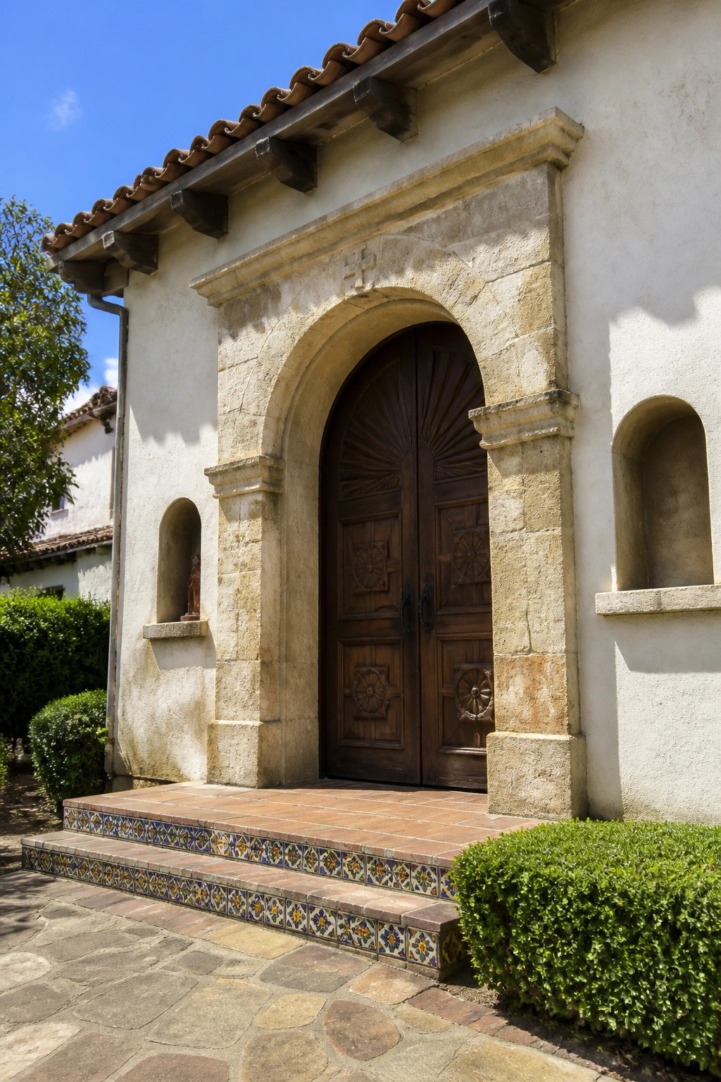 A front entrance featuring double wooden doors under a stone arch, with tiled steps and recessed niches on either side of the doorway.
