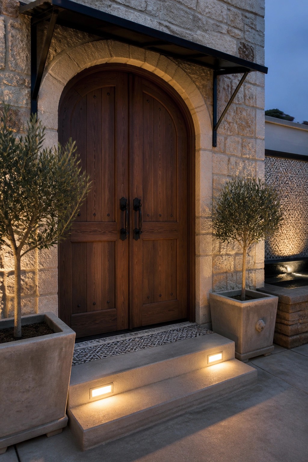 A double arched wooden door set in a light stone arch above three concrete steps with recessed lights, flanked by two potted trees.