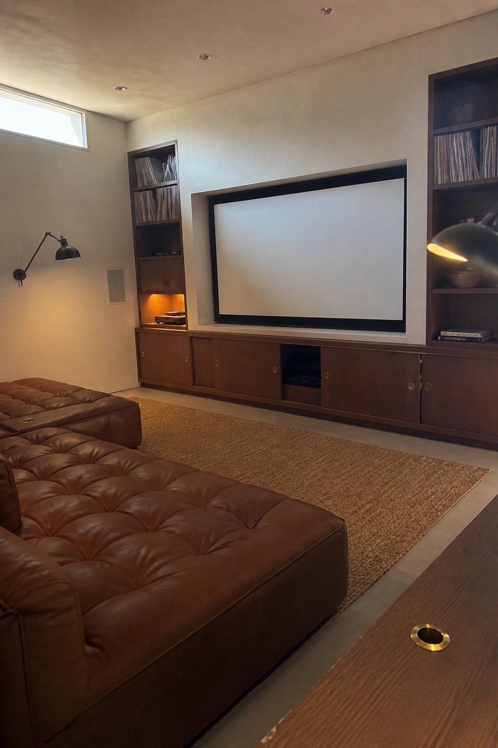 A modern home cinema with a large screen centered on a light wall, flanked by dark wood built-in shelves and cabinets holding vinyl records and AV equipment, and a brown leather sectional sofa on a woven rug in the foreground.