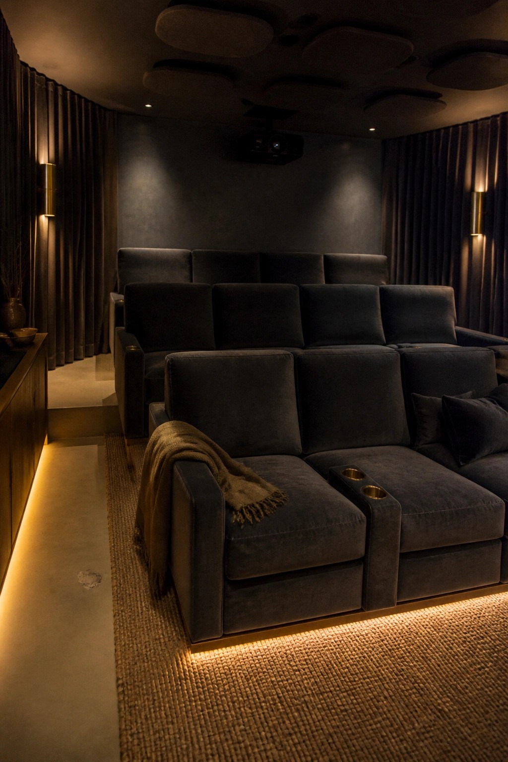 A home theater with three rows of dark gray velvet seating on tiered platforms, a ceiling-mounted projector, wall sconces, and side curtains.