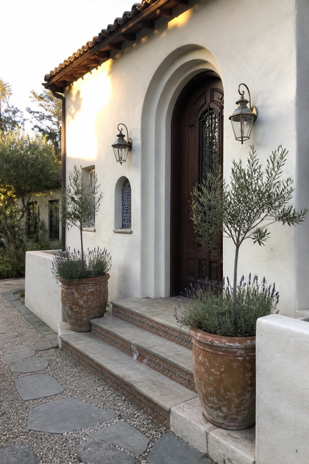 White stucco entrance with arched wooden door, lanterns, and potted lavender on tiled steps.