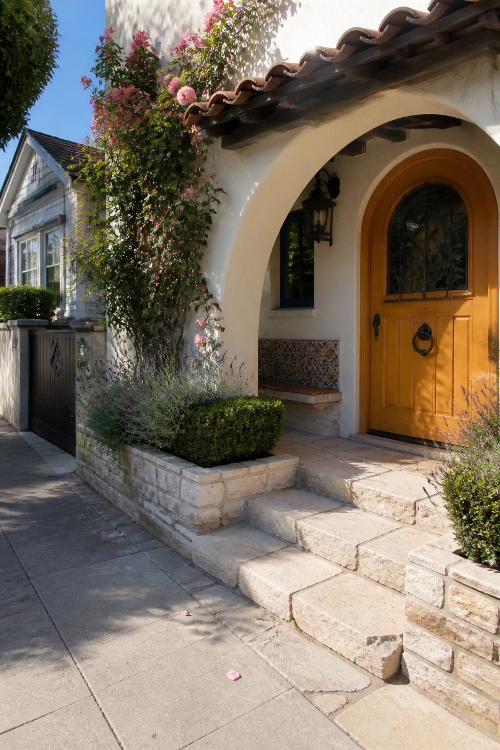 A Mediterranean-style home entrance with a bright yellow wooden door inside a white stucco arch, stone steps, and climbing pink roses on the wall.