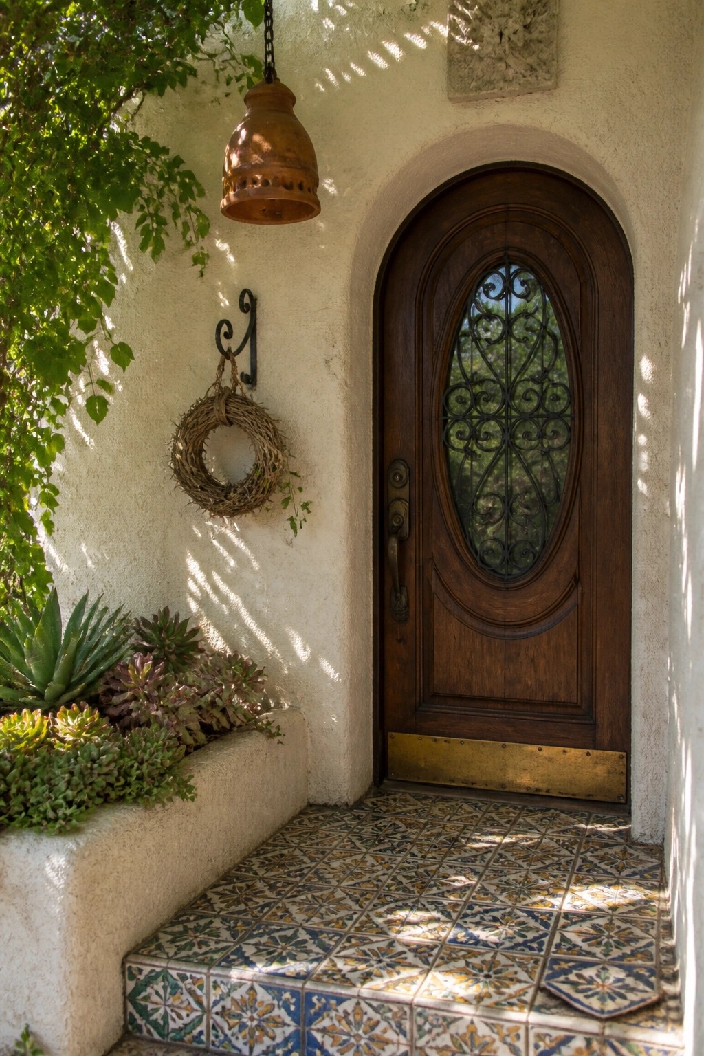 A dark wooden front door with an oval iron grille inside an arched stucco entry above patterned tile steps.