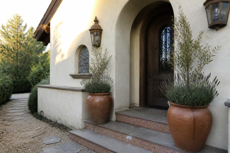 22 Architectural Luxury Front Doors for Mediterranean-Style Homes White stucco entrance with arched wooden door, lanterns, and potted lavender on tiled steps.