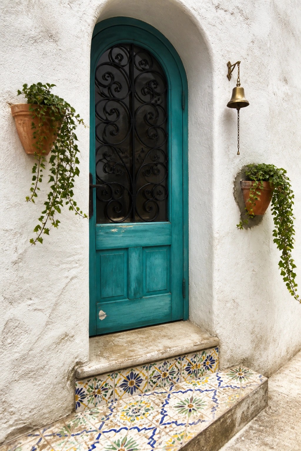 A turquoise arched door with black wrought iron detailing in a white stucco wall, featuring colorful patterned tiles on the steps and two terracotta pots with trailing plants.