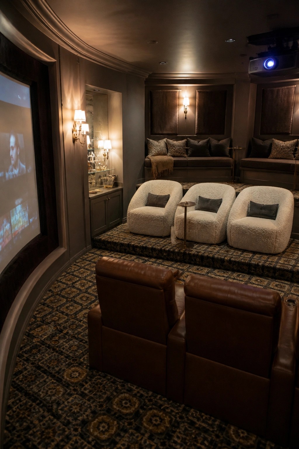A dimly lit home cinema room showing tiered seating with brown leather recliners in front and white upholstered chairs on a raised carpeted platform facing a projection screen.