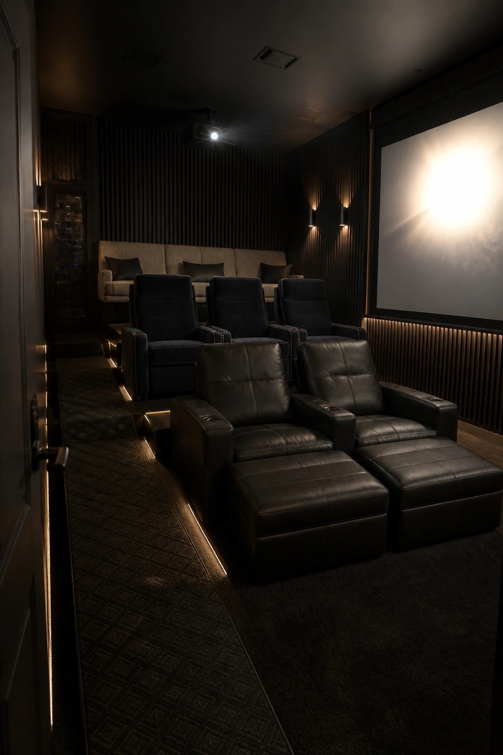 A dimly lit home cinema with several rows of black leather recliners arranged on tiered platforms facing a large projection screen.