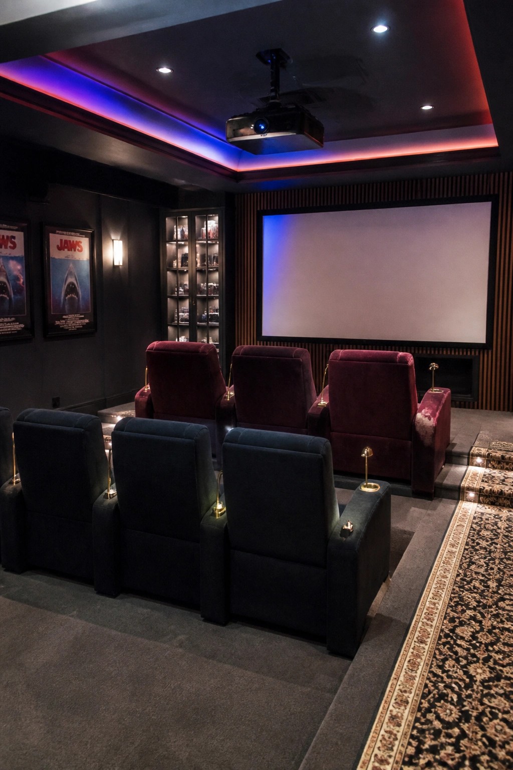 A home cinema room with two rows of dark blue recliners in front and three rows of red velvet recliners behind them on a raised platform, facing a large projection screen with a ceiling-mounted projector above.