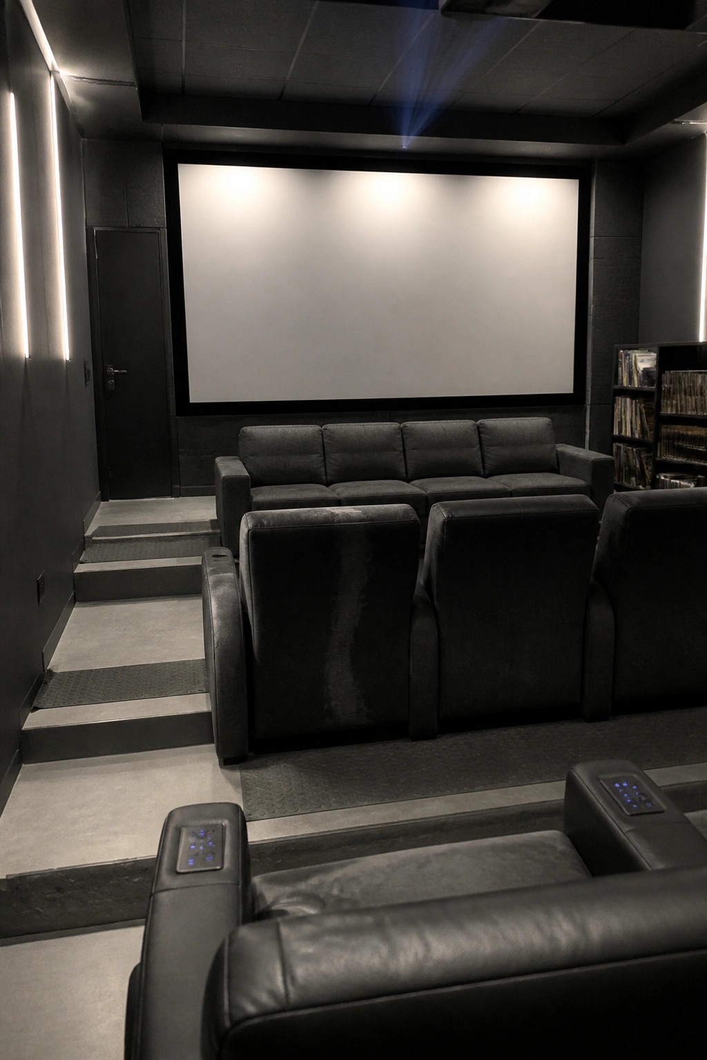 A dark home cinema room with multiple rows of black leather recliners arranged on tiered platforms facing a large blank screen.