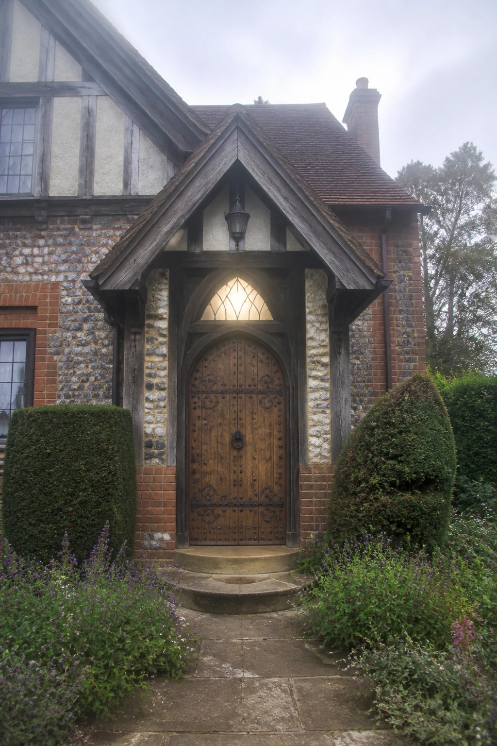 A large wooden arched front door with black metal hardware and a pointed transom window sits beneath a steep wooden gable porch on a brick and flint Tudor house.